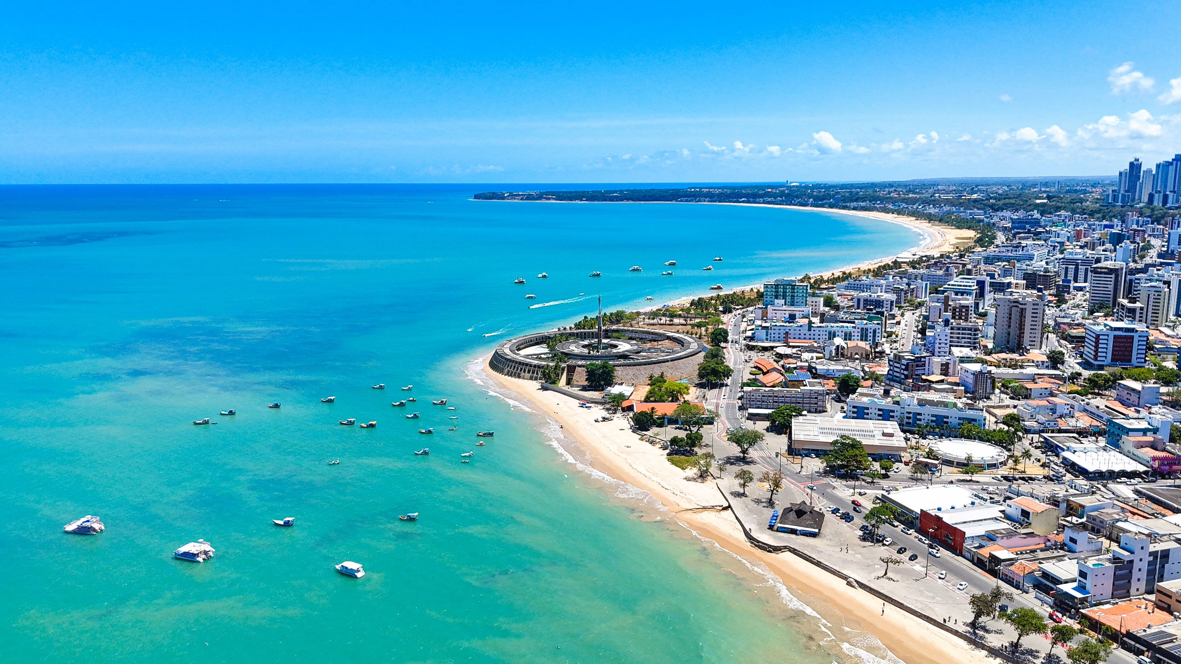 Vista aérea da orla de João Pessoa, com a praia, os barcos e a cidade acompanhando a costa